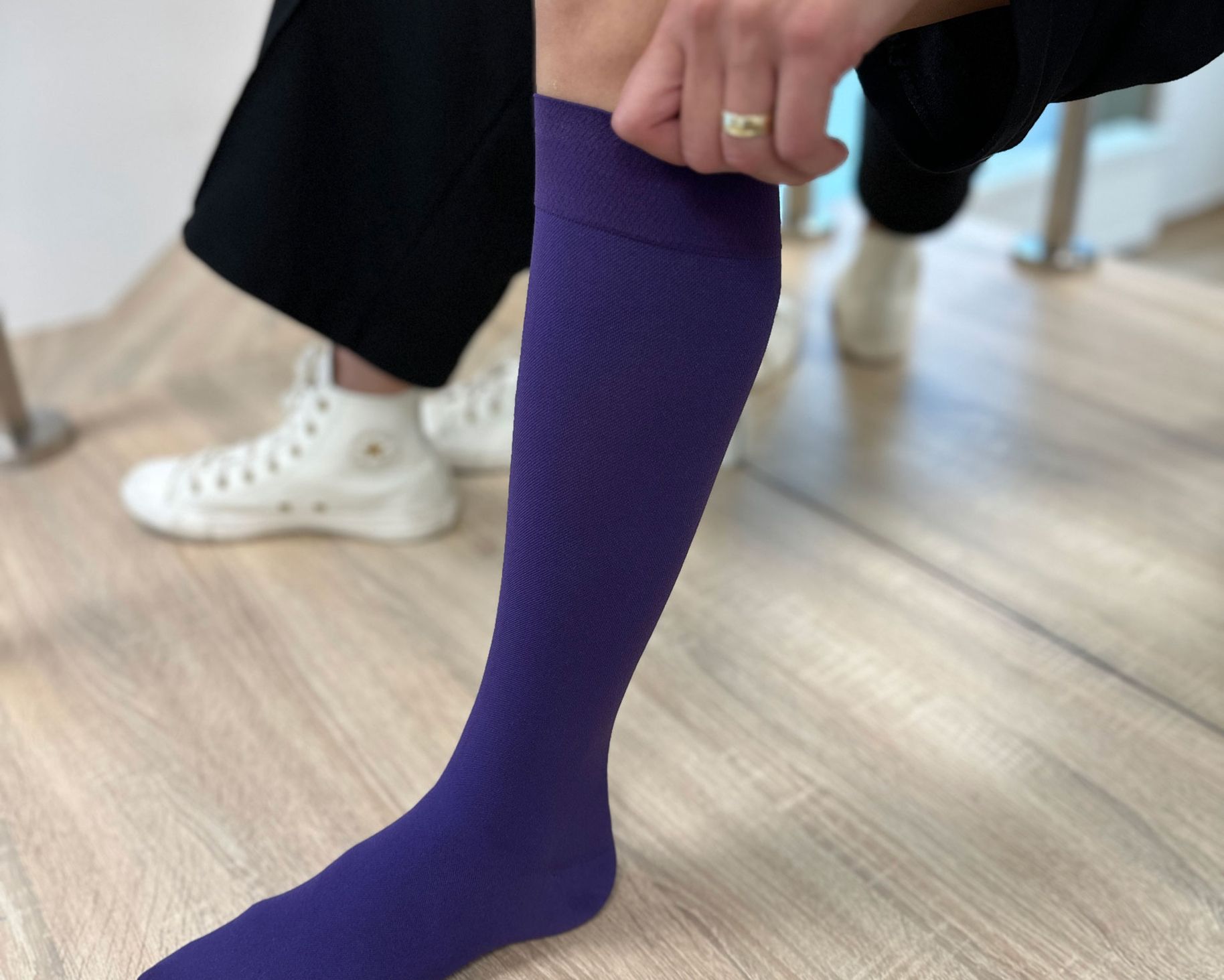 Eine Person zieht einen lila Kniestrumpf an. Im Hintergrund sind eine andere Person in schwarzen Hosen und weiße Sneakers zu sehen.