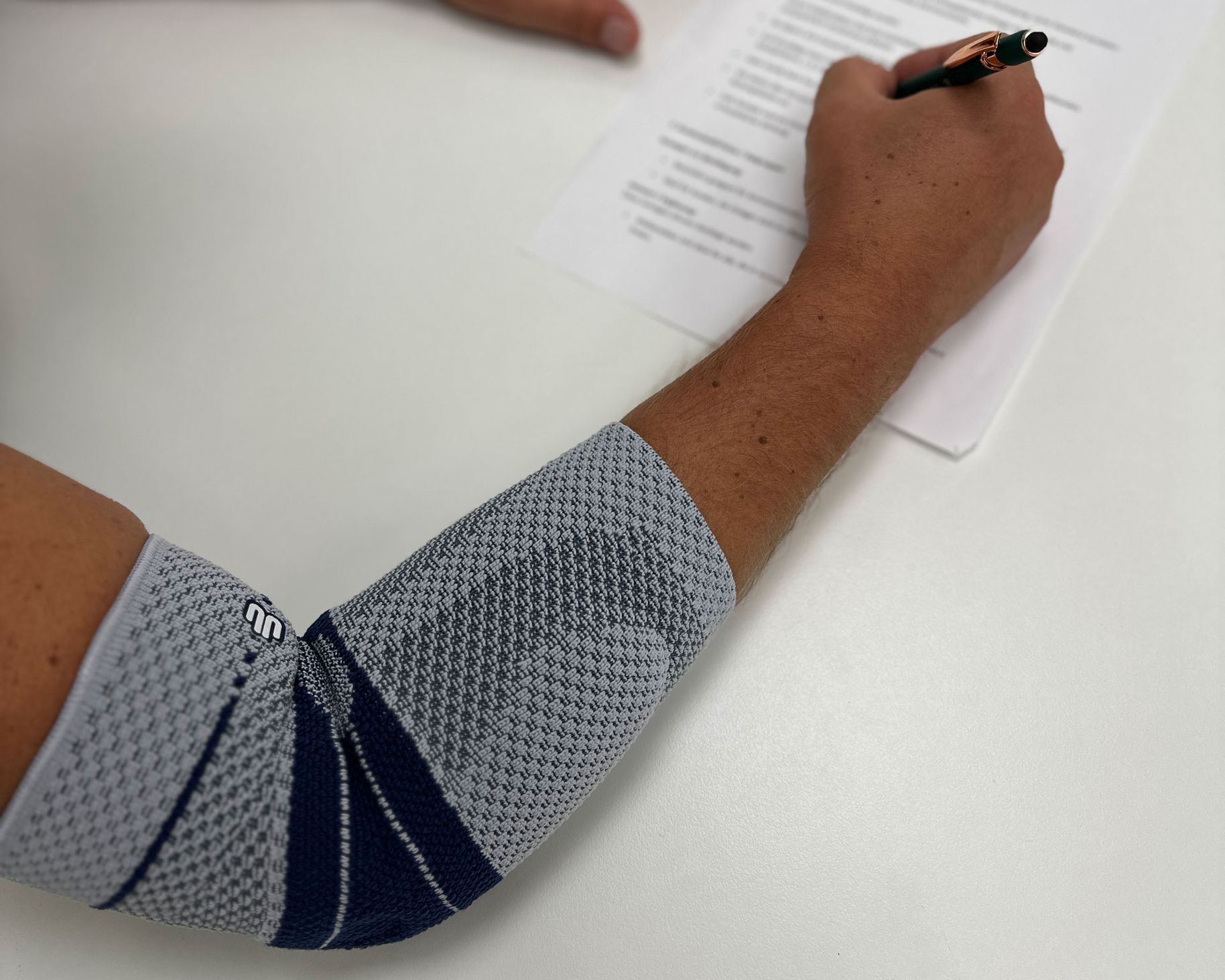 Ein menschlicher Arm mit einem grauen, elastischen Armstützverband, der eine Hand beim Schreiben auf einem Blatt Papier unterstützt.