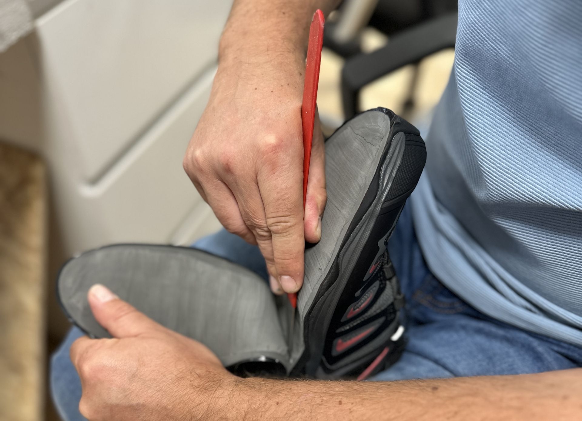 Person in Jeans und blauem T-Shirt hält eine graue Sportschuhsohle und bearbeitet sie mit einem roten Werkzeug