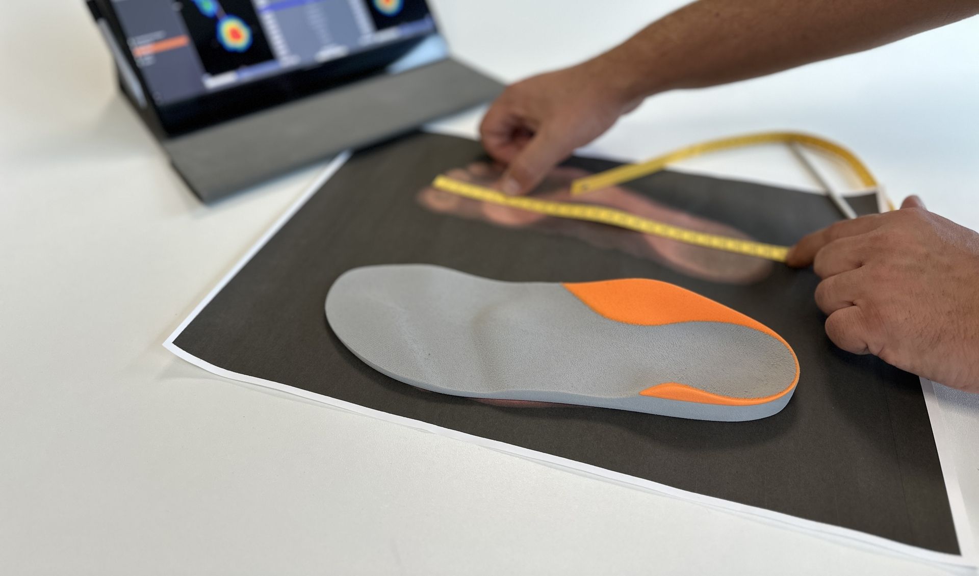 Hand misst mit gelbem Maßband eine graue Einlegesohle mit orangefarbenen Flächen auf einem schwarzen Blatt, im Hintergrund ein Tablet mit Fußdruckmessung