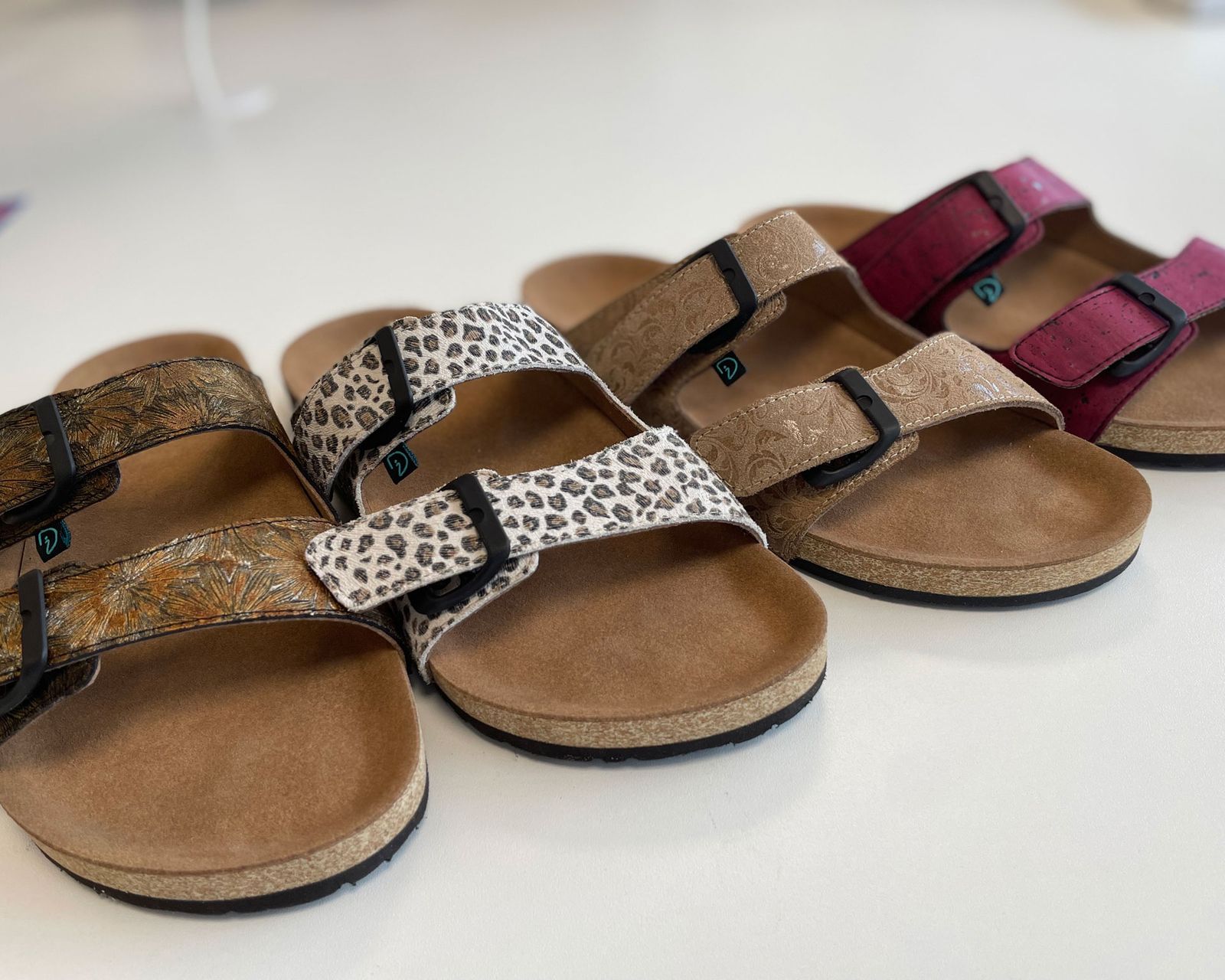 Vier Paar Sandalen in verschiedenen Designs: ein Paar mit goldenen Mustern, ein Paar mit Leopardenmuster, ein Paar in Beige und ein Paar in Pink. Alle haben eine Korksohle und schwarze Riemen.