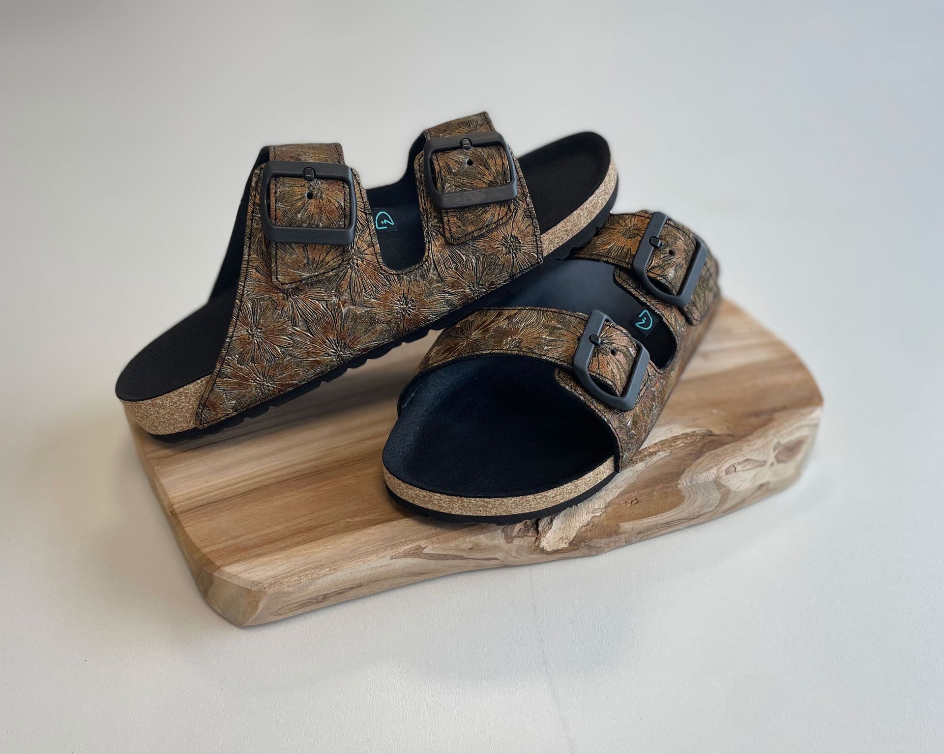 Braune Sandalen mit zwei Schnallen auf einer Holzplatte. Die Sandalen haben ein florales Muster und eine schwarze Innensohle.