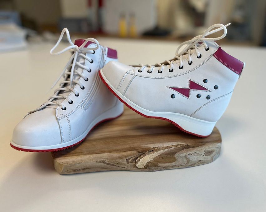 Weiße Sportschuhe mit pinken Akzenten, hoher Form und Schnürung, auf einem Holzbrett platziert.