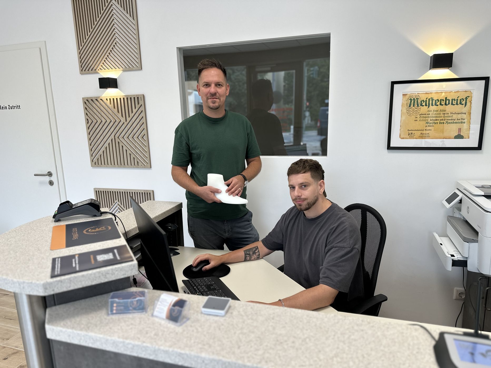 Zwei Männer an einem Empfangstresen, einer sitzt am Computer, der andere steht mit einem Papier in der Hand, im Hintergrund ein Meisterbrief an der Wand.