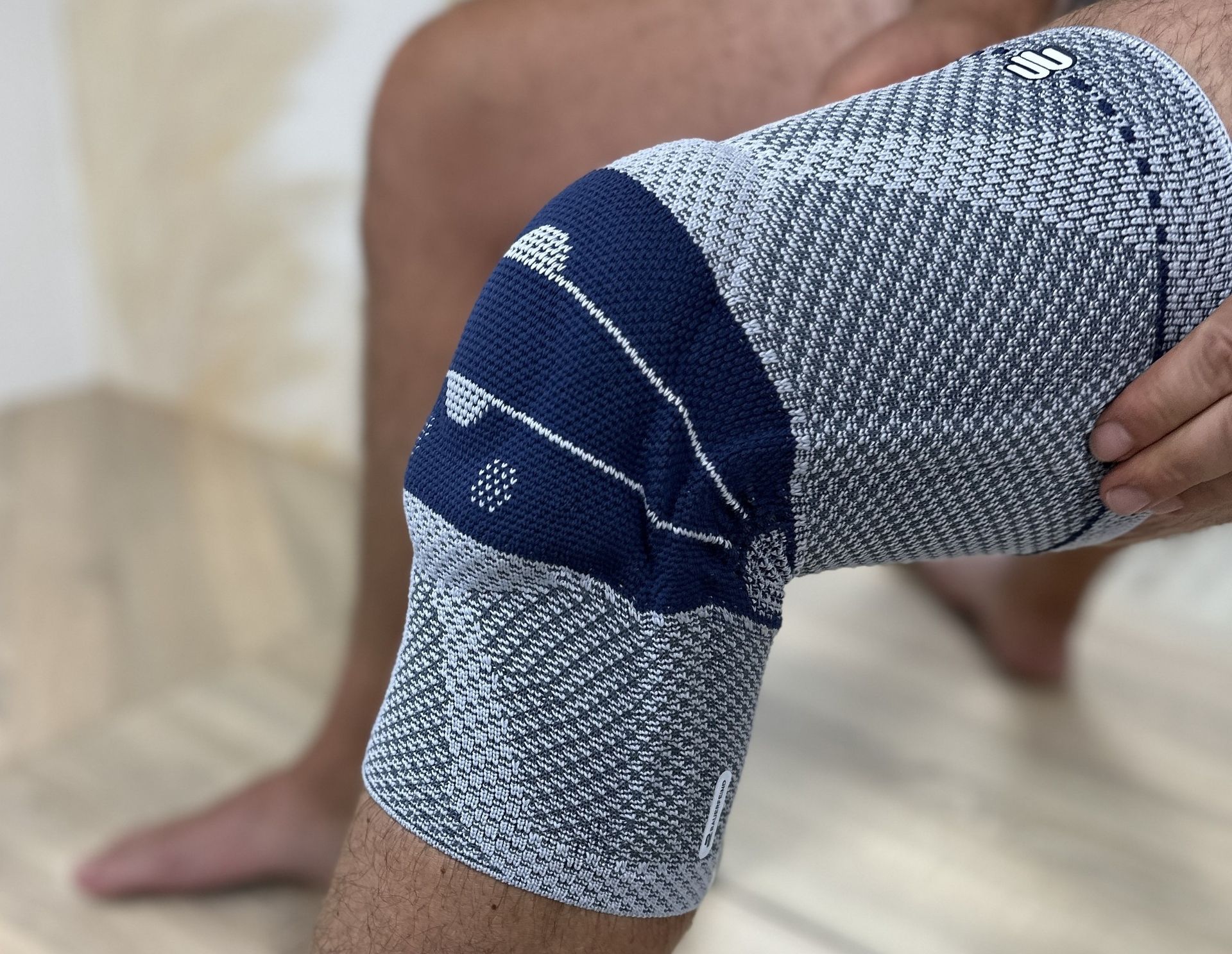 Bein mit grauer und dunkelblauer Kniebandage, Hand hält das Knie, Holzboden im Hintergrund