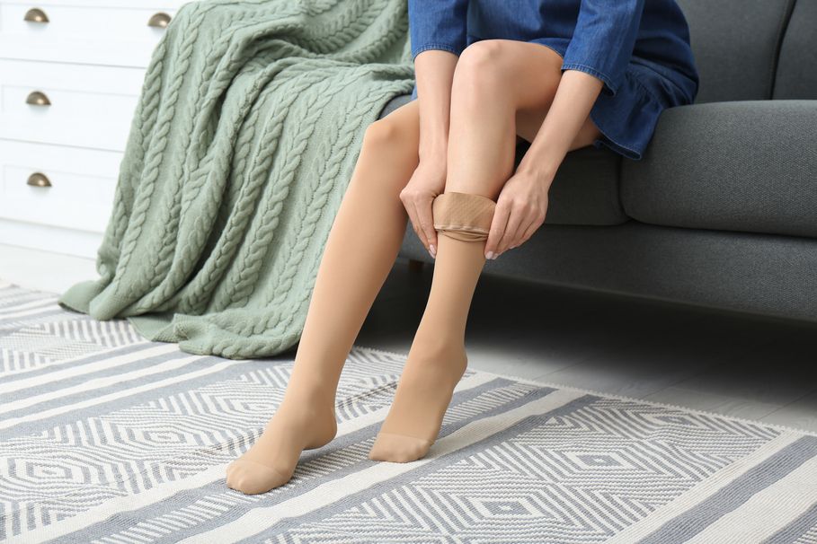 Eine Person sitzt auf einem grauen Sofa und zieht einen beigen Kompressionsstrumpf über das Bein. Im Hintergrund liegt eine grüne Decke und der Boden ist mit einem gemusterten Teppich bedeckt.