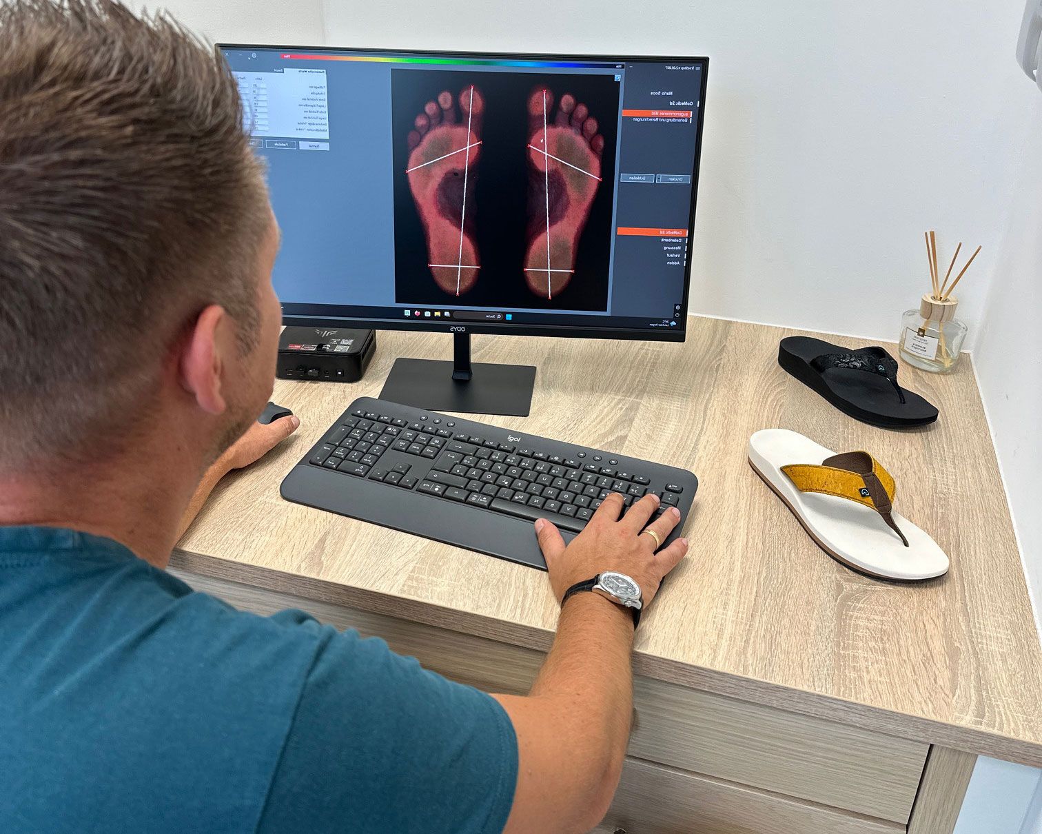 Mann mit kurzen Haaren sitzt vor einem Computer, der ein Fußscan-Bild zeigt. Auf dem Tisch liegen ein schwarzer Schuh und eine Sandale mit gelbem Riemen.