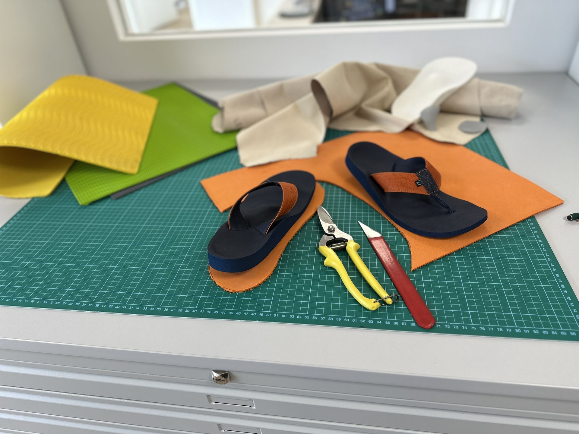 Zwei dunkelblaue Sandalen mit braunen Riemen auf orangefarbenem Leder, daneben gelbe Zangen und ein rotes Messer auf grünem Schneidbrett, im Hintergrund verschiedene Lederstücke und ein beige-farbenes Kleidungsstück.