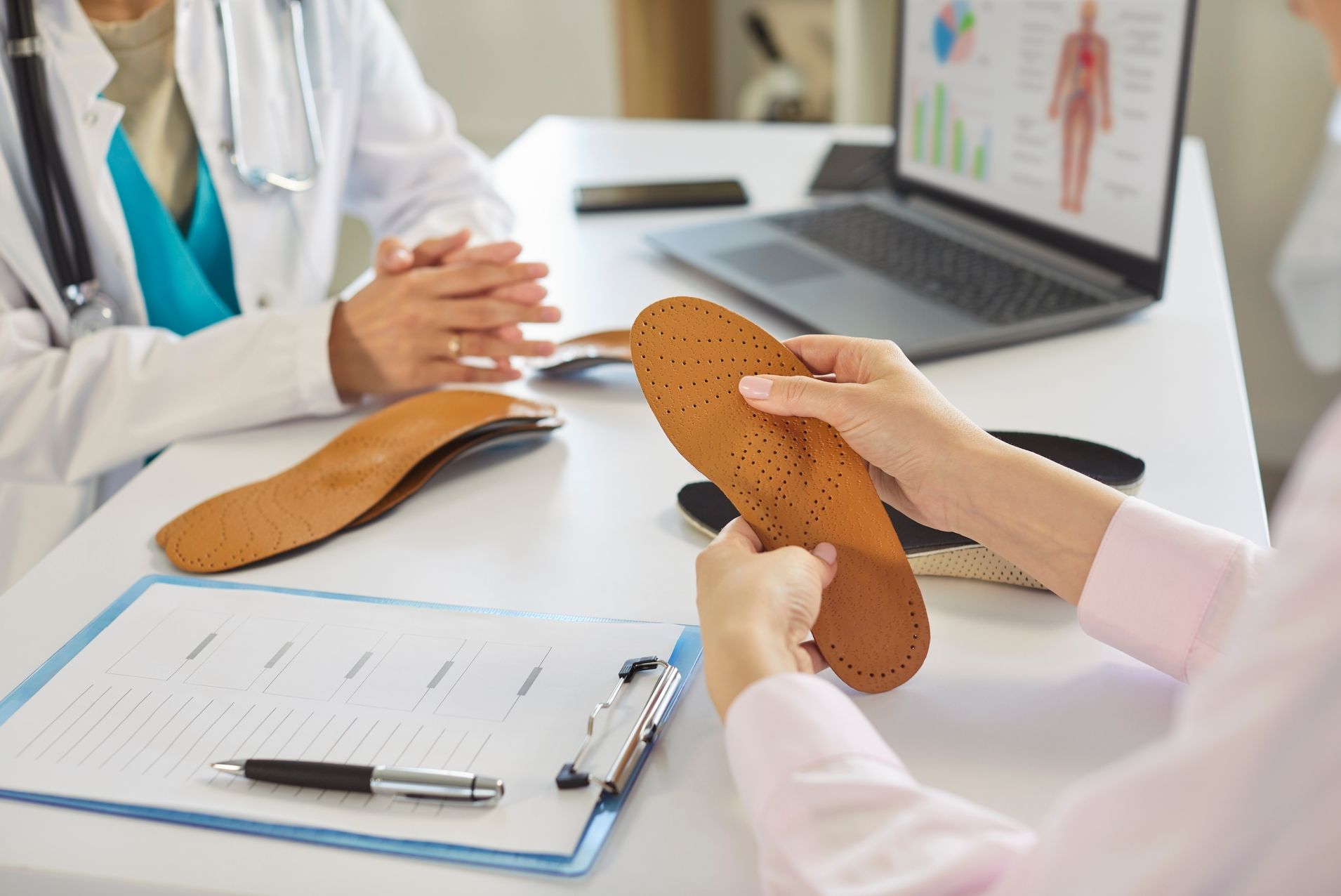 Person hält braune, perforierte Schuheinlage, Arzt im Hintergrund mit verschränkten Händen, Schreibtisch mit Klemmbrett und Laptop mit medizinischer Grafik