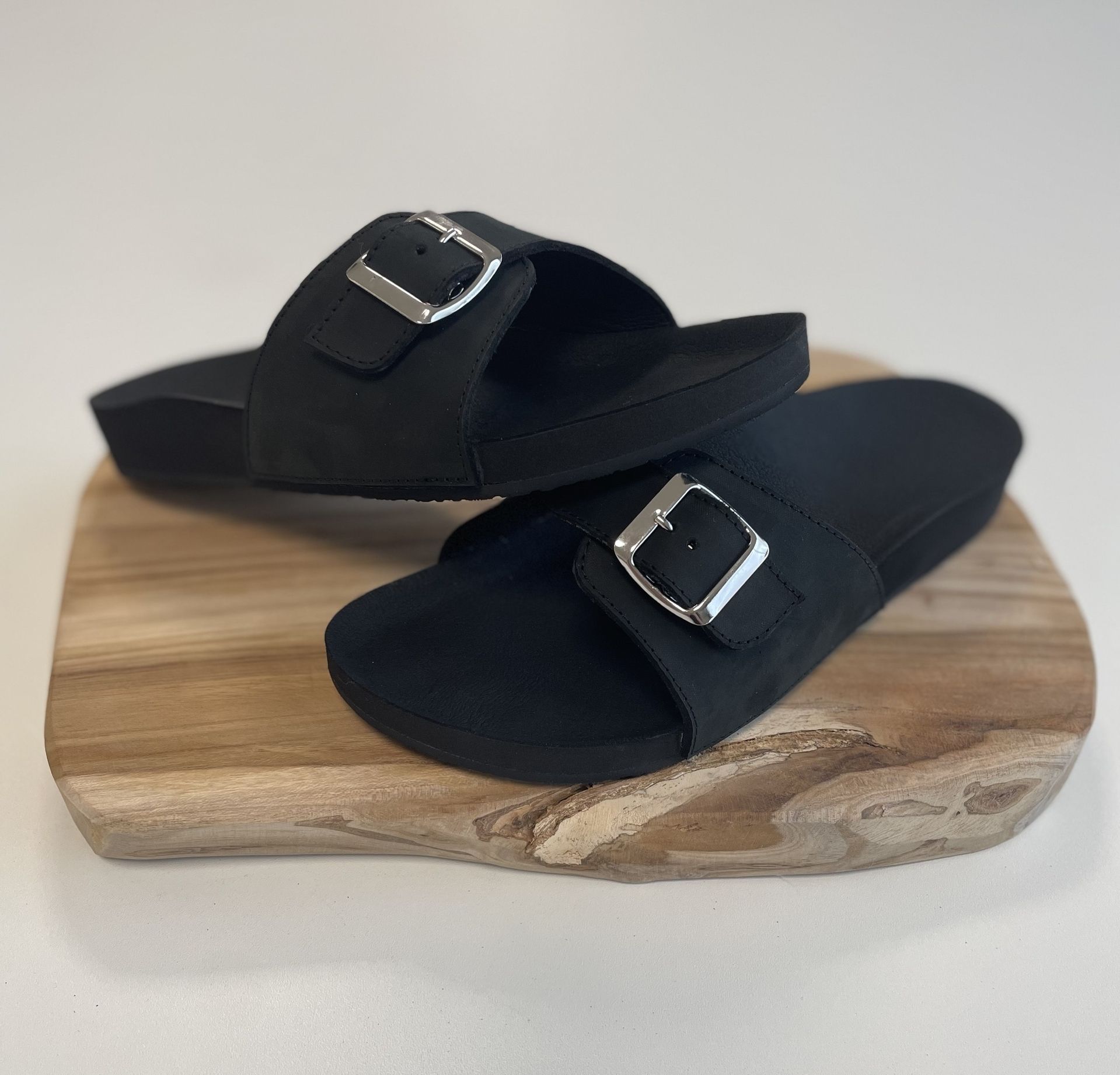 Schwarze Sandalen mit breitem Riemen und silberner Schnalle auf Holzbrett